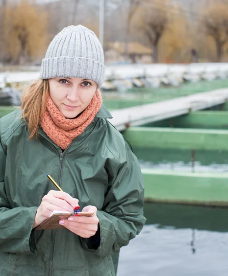 Technicienne aquacole prenant des notes sur un site de pisciculture en extérieur, illustrant le suivi en nutrition animale.