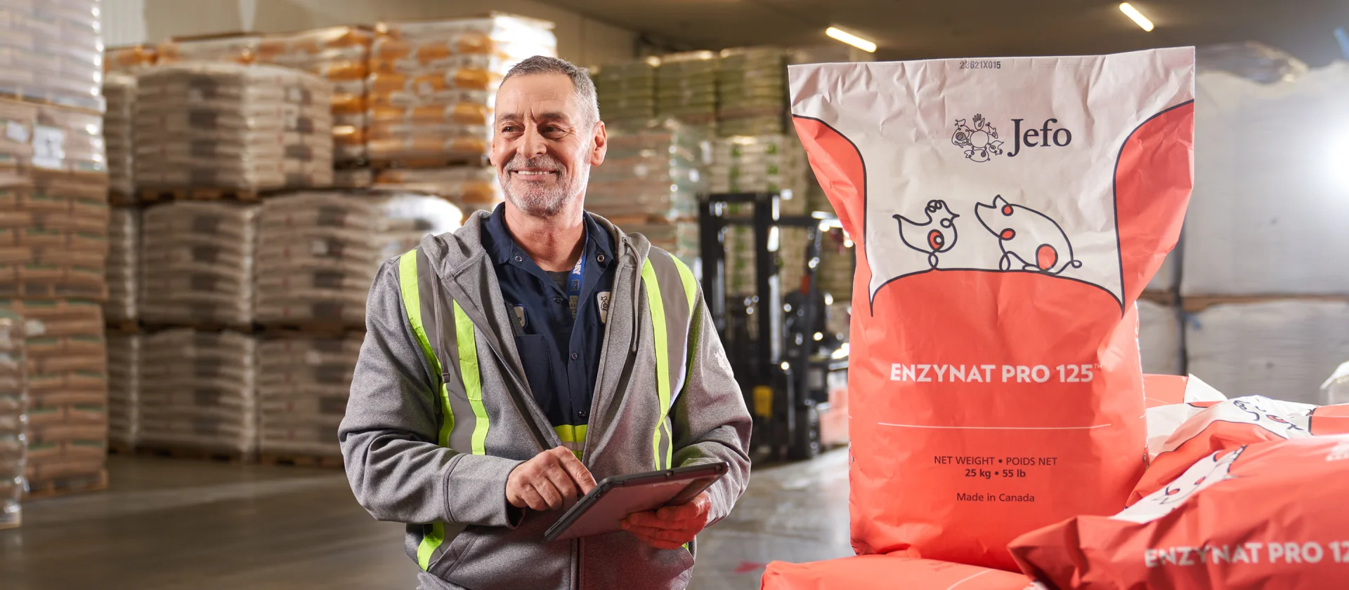 Jefo employee in warehouse using a tablet beside Enzynat Pro 125 feed bag for swine and poultry animal nutrition.